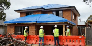 Construction workers in PPE erecting temporary roof coverings and safety barriers around a storm damaged residential building in Sydney